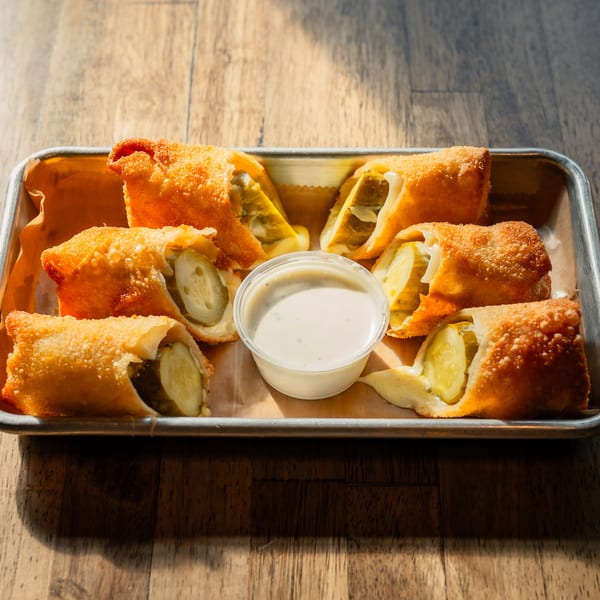 Golden-fried pickle egg rolls with melted cheese and a side of ranch dressing on a metal tray.