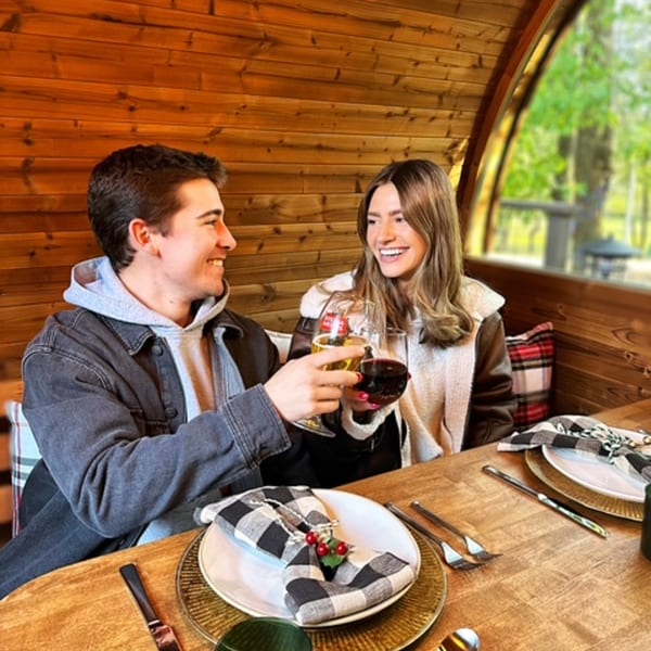 A smiling couple toasts drinks inside a cozy wooden dining pod at Magpie's Den & Pen.