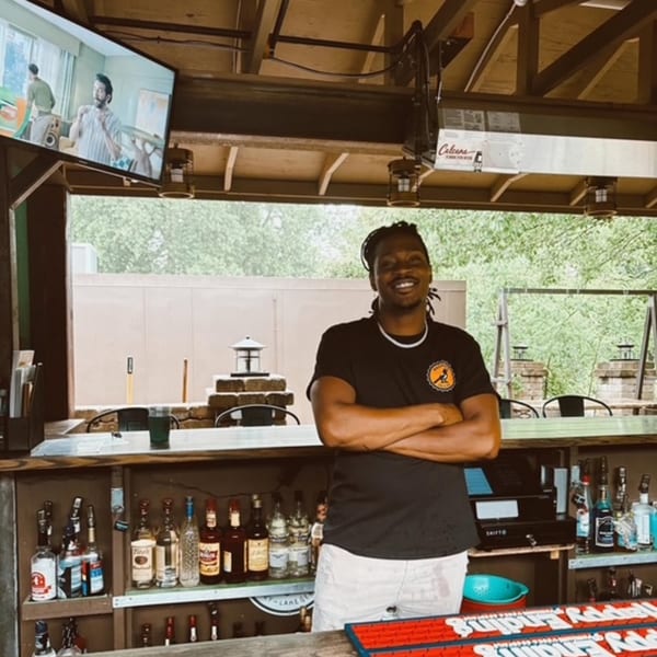 Smiling bartender with dreadlocks and a Magpie's Den & Pen shirt stands behind a bar.