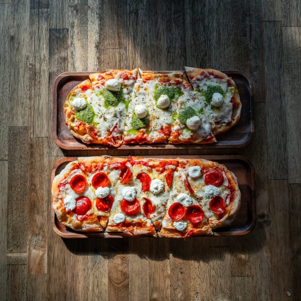 Two rectangular flatbread pizzas, one pesto and one pepperoni with ricotta, on wooden trays.