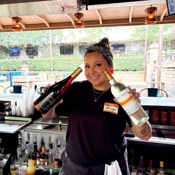 A smiling bartender named Maggie holds up two bottles of wine behind a bar at Magpie's Den & Pen.