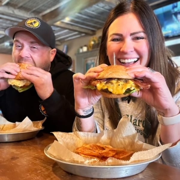 Two happy customers at Magpie's Den & Pen enjoying large burgers with sides of chips.