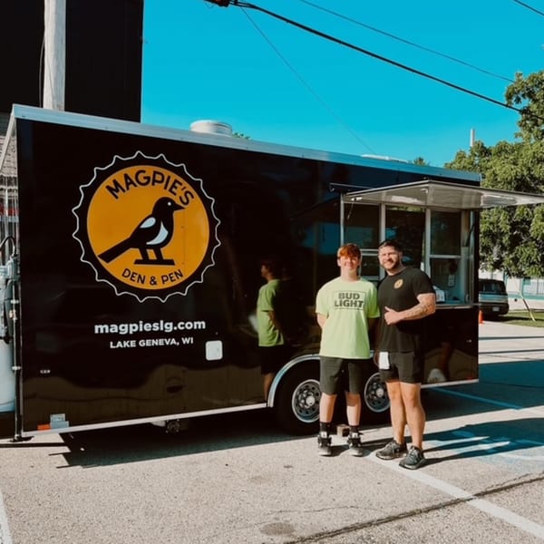 Two men stand proudly in front of the black Magpie's Den & Pen food truck on a sunny day.
