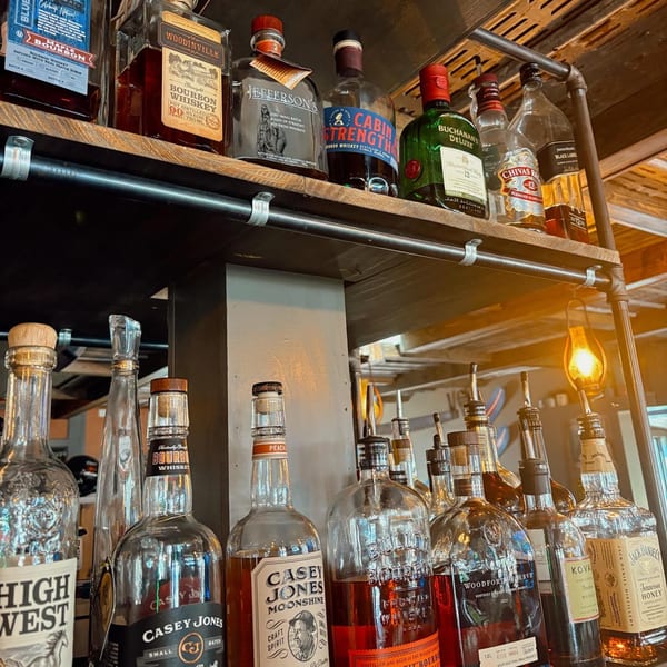 A bar shelf at Magpie's Den & Pen filled with various bottles of bourbon, whiskey, and moonshine.